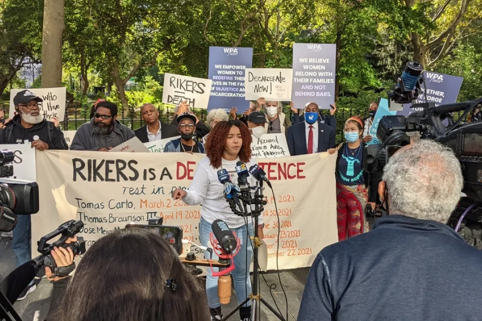 Crowd of activists protesting conditions at Rikers Island with one woman in front speaking into microphone